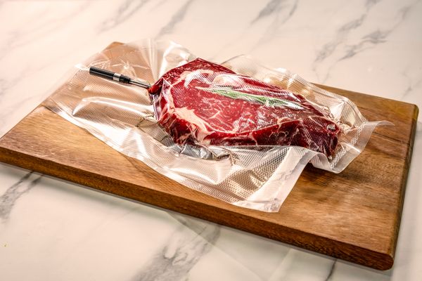 A vacuum-sealed steak with a sprig of rosemary and a meatprobe inserted, resting on a wooden cutting board.