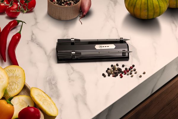 A sleek NEFF meatprobe rests on a marble countertop surrounded by vibrant vegetables and mixed peppercorns.