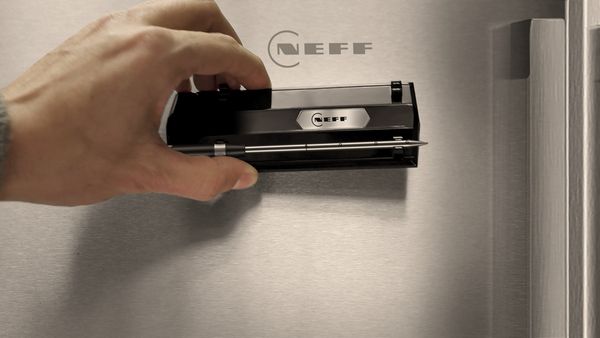 A hand holds a black NEFF magnetic meatprobe against a stainless steel refrigerator surface.