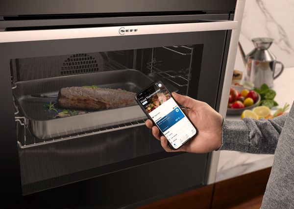 En hånd holder en smarttelefon som viser matlagingsinformasjon, mens en laksefilet tilberedes i en Neff-ovn.