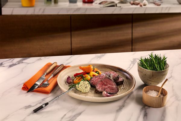 A plate of perfectly cooked beef with vegetables and herb butter on a marble countertop.