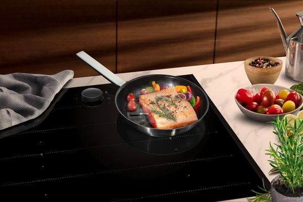 A frying pan with salmon and colorful vegetables sizzles on a sleek black induction hob. 