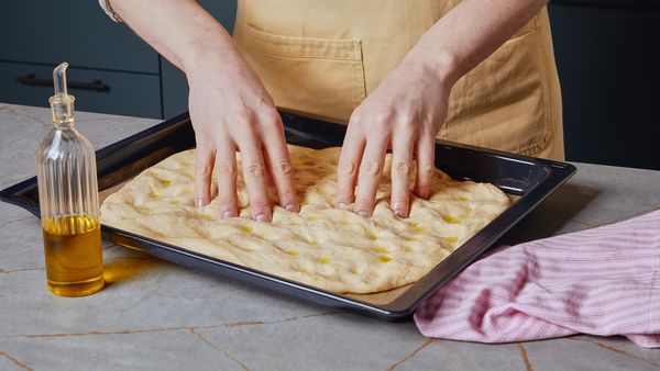 Zwei Hände drücken Vertiefungen in einen ausgerollten Teig, der mit Öl bestrichen wurde.