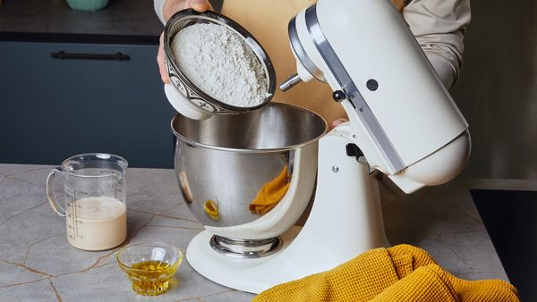 Eine Person gibt Mehl aus einer Schüssel in die Rührschüssel einer Küchenmaschine.