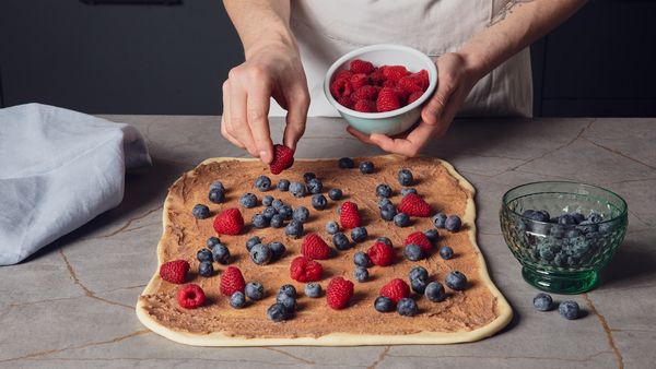 Eine Hand legt Himbeeren auf einen ausgerollten Teig mit brauner Creme auf einer Küchenarbeitsplatte.