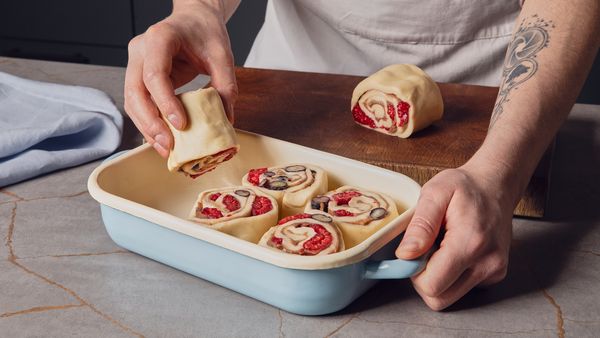 Eine Hand legt ungebackene Zimtbrötchen, aus denen Beeren hervorblitzen, in eine rechteckige blaue Backform auf einer Küchenarbeitsplatte.