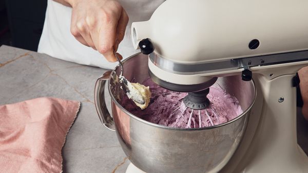 Person adding a spoon of butter to the bowl of a stand mixer which is filled with a lilac cream