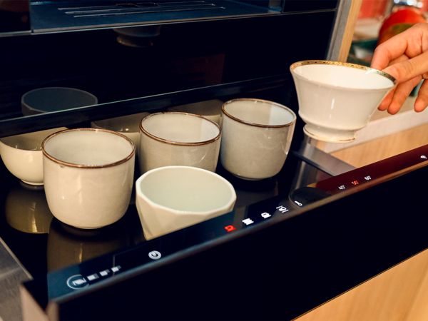 Близък план на ръка, държаща декоративна чаша за чай, над няколко обикновени бели и сиви чаши в чекмедже за затопляне.