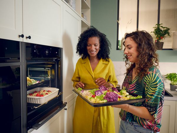 Two women stand in a kitchen, holding trays of food in front of an open oven, smiling at each other. 