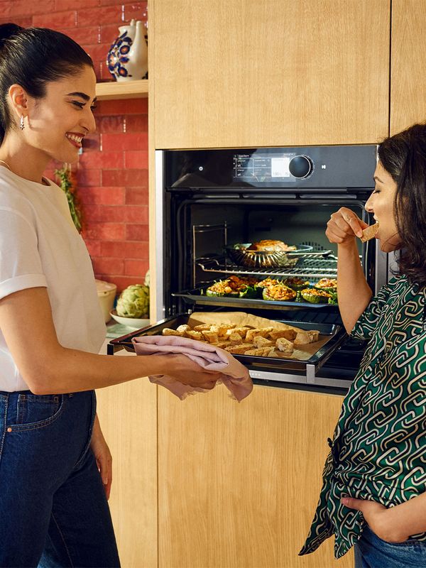 Two women stand together in front of an oven, engaged in conversation while preparing a meal. 