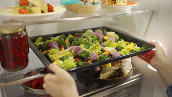 Una mano afferra una teglia di verdure miste dall'aspetto invitante, con cavolfiori viola e verdi, all'interno di un frigo ben fornito.
