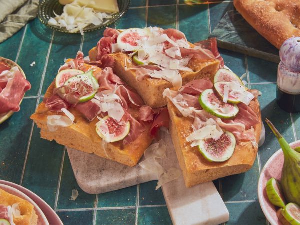 Drei Stücke Foccacia mit Schinken, Käse und Feigenspalten in der Bildmitte auf blauen Küchenfliesen, umgeben von Geschirr.