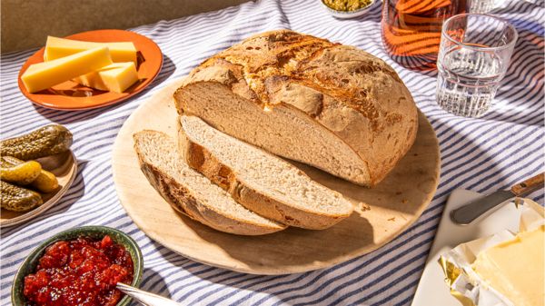 Handgemachtes Brot auf einer Holzservierplatte mit Käse, Pickles, Marmelade und Butter, auf einer gestreiften Tischdecke.