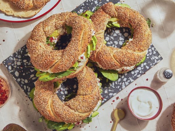 Drei Simits liegen auf einem schwarz-weißen Steinbrett auf einer weißen Tischdecke, umgeben von Geschirr.