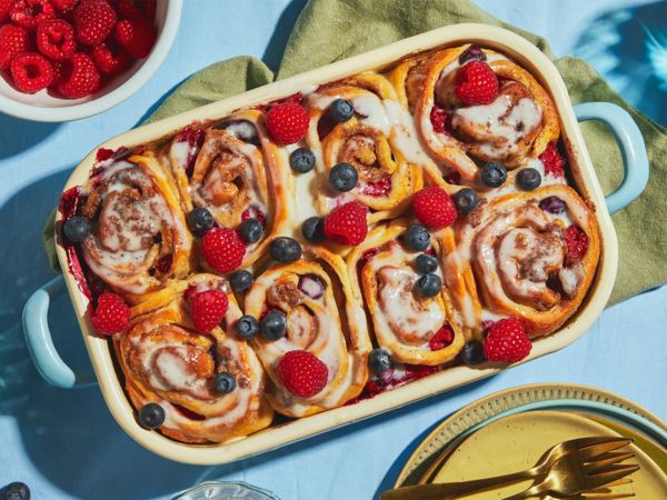 Gebackene Zimtbrötchen mit frischen Beeren und weißem Frosting in einer rechteckigen Backform stehen auf grünen und blauen Tischdecken, umgeben von Geschirr.