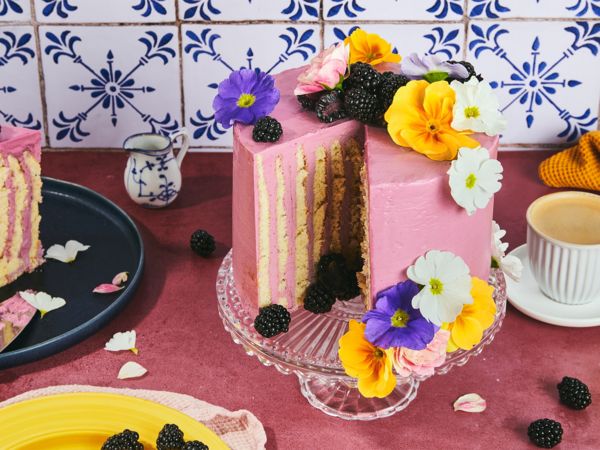 A blackberry stripe cake with vertical layers and a piece cut out of it sits on a glass cake stand placed on a pink table
