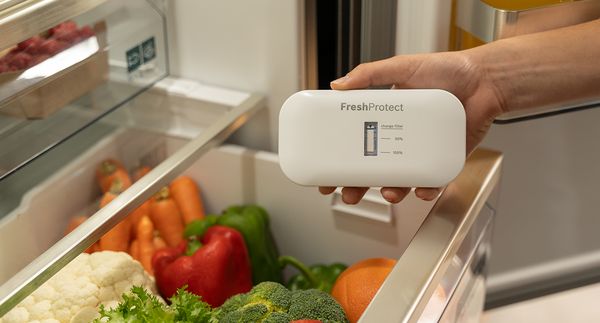 Main tenant le dispositif FreshProtect au-dessus de légumes colorés dans un réfrigérateur pour optimiser leur fraîcheur.