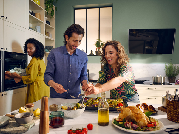 Three people prepare and serve food in a modern kitchen; one person puts a tray in the oven while two plate roasted vegetables at the counter.