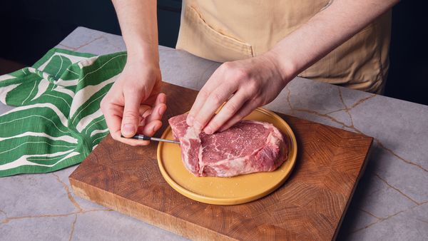 Eine Hand steckt ein kleines Fleischthermometer in die Seite eines rohen Steaks, das auf einem Holzschneidebrett liegt.