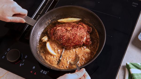 Ein Steak wird in einer schwarzen Pfanne zusammen mit einer Sauce und Zitronenspalten gegrillt.