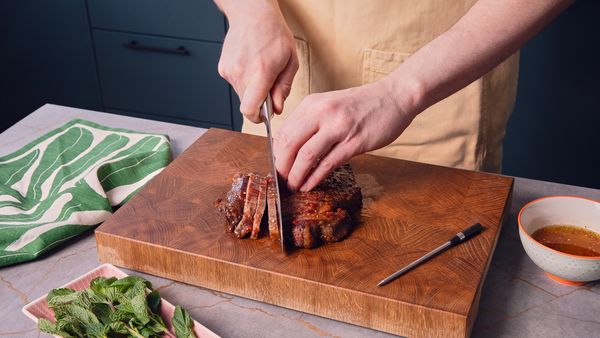 Zwei Hände schneiden ein gegrilltes Steak auf einem Holzschneidebrett in Streifen.