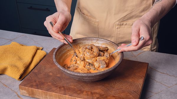 Zwei Hände vermengen Garnelen und eine braune Sauce in einer Schüssel, die auf einem Schneidebrett steht.