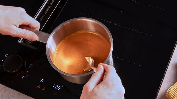 Eine Hand hält einen mit Sauce bedeckten Teelöffel über einem Kochtopf mit der Sauce auf dem Kochfeld.