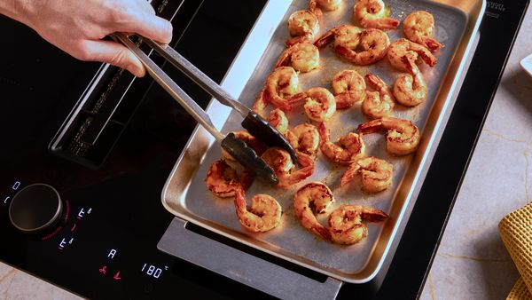 Eine Hand wendet mit einer Zange Garnelen, die auf einer silberfarbenen Grillplatte gegrillt werden.