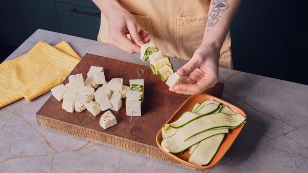 Two hands are putting zucchini slices and halloumi cubes onto skewers over a wooden cutting board.