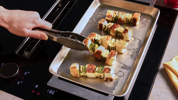 A hand is turning halloumi zucchini skewers on a silver grilling plate with a pair of tongs.