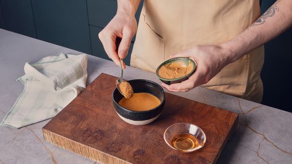 Eine Hand füllt einen Löffel Erdnussbutter in eine Schüssel, die zur Hälfte mit Flüssigkeit gefüllt ist.