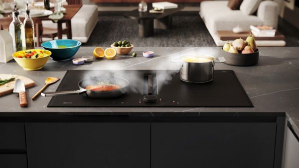 Een inductiekookplaat met pannen waarin gekookt wordt, omringd door verse ingrediënten. De stoom van het eten wordt weggezogen door de geïntegreerde afzuiging.