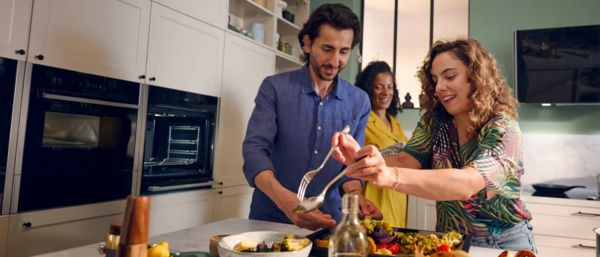 Zwei Frauen halten in einer K&uuml;che ein mit Gem&uuml;se gef&uuml;lltes Backblech, neben einem roten Kreis mit deutschem Text &uuml;ber ein 100&euro; Cashback-Angebot.