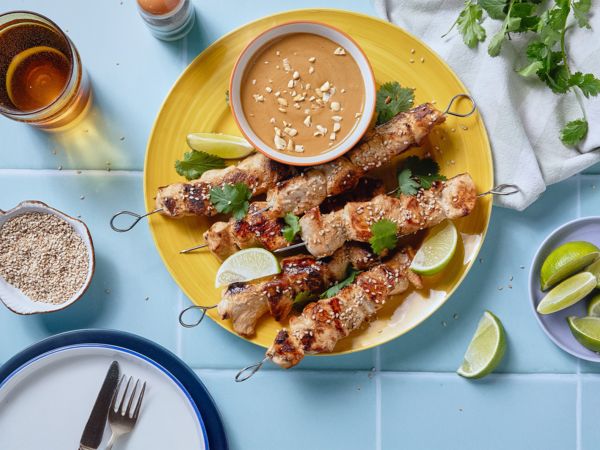 Gebratene Hähnchenstücke auf Grillspießen auf einem gelben Teller, daneben eine Schüssel mit einem braunen Dip. 