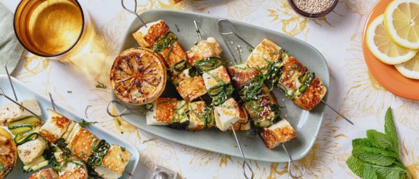 Fried halloumi pieces on metal skewers lay on a blue rectangular plate surrounded by other plates and tableware.