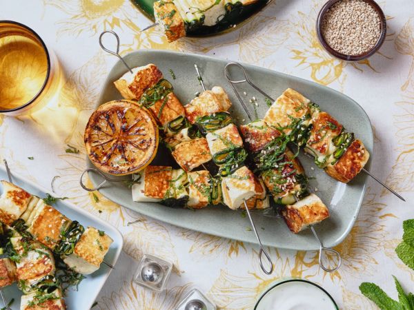 Halloumi fritto su spiedi di metallo disposti su un piatto rettangolare blu, circondato da altri piatti e stoviglie.