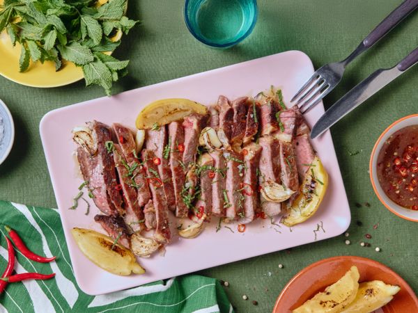 Ein gegrilltes und in Streifen geschnittenes Steak liegt auf einem rechteckigen, rosafarbenen Teller, der auf einer grünen Tischdecke steht, umgeben von Geschirr. 