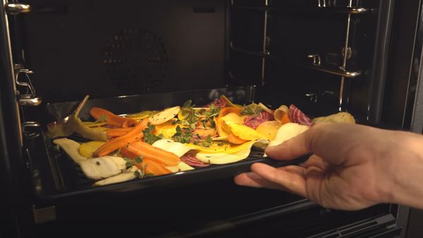 Ръка вкарва във фурна тава с тънко нарязани на филийки зеленчуци, гарнирани с билки и зехтин.