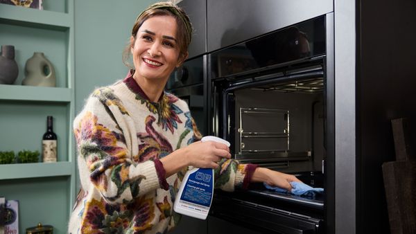 Sieviete raibā blūzē stāv ar tīrīšanas līdzekļa pudeli un tīra krāsni iekšpuses.