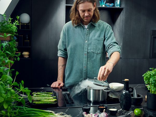 Uomo che cucina su un piano cottura a induzione con cappa integrata Neff, mentre solleva il coperchio di una pentola fumante