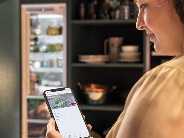 Una persona tiene in mano uno smartphone con un'app aperta che mostra il frigorifero, con una cucina arredata in stile moderno sullo sfondo e un frigorifero aperto con tante pietanze al suo interno.