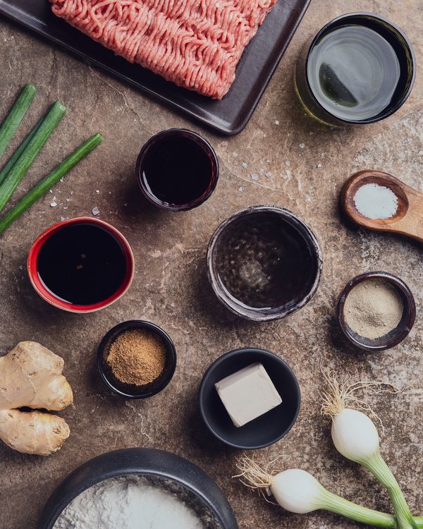 Spread of ingredients for steamed pork buns including ground meat, scallions, ginger, flour, spices, sauces, yeast, and water arranged on a kitchen counter.