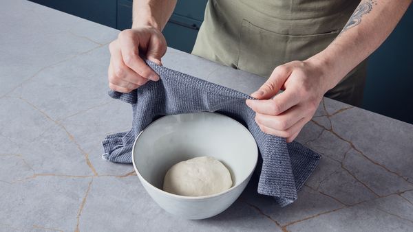 Person covering a bowl of dough with a towel on a kitchen counter, preparing the dough to rise.