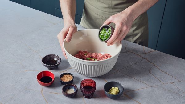 Person adding chopped scallions into a bowl of ground meat surrounded by small bowls of sauces and minced ginger for the pork bun filling.