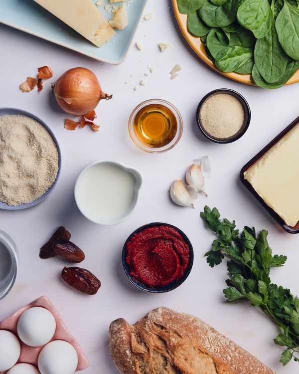 Spread of ingredients including spinach, eggs, parmesan, onion, bread, garlic, dates, tomato paste, milk, parsley, and butter arranged on a light surface.