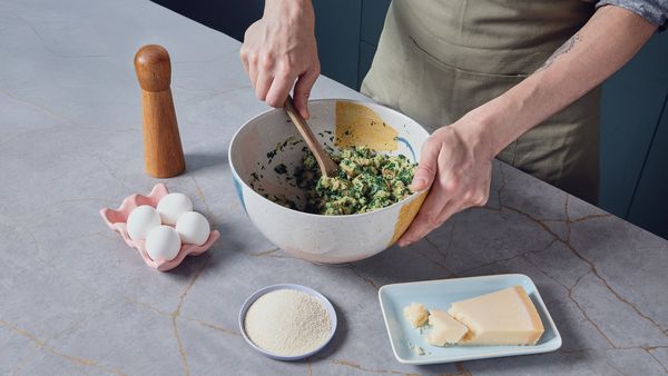 Person mixing chopped spinach, bread, and cheese in a bowl with a wooden spoon, surrounded by eggs, pepper mill, grated cheese, and breadcrumbs.
