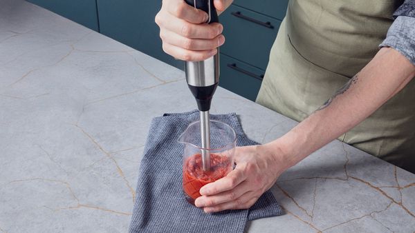 Person blending tomato-date sauce with an immersion blender in a measuring cup placed on a cloth-covered kitchen counter.