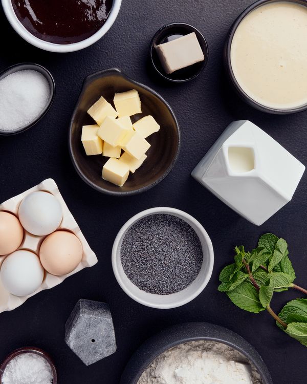 Sprerad of ingredients including butter cubes, eggs, poppy seeds, flour, yeast, sugar, milk, jam, and fresh mint on a black surface.