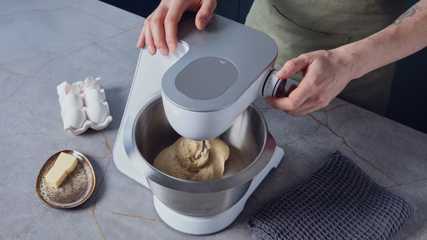 Person using a stand mixer to knead dough, with eggs and butter nearby on the kitchen counter.