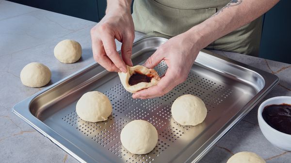 Person stuffing dough with plum jam to shape dumplings on a perforated tray, ready for steaming.